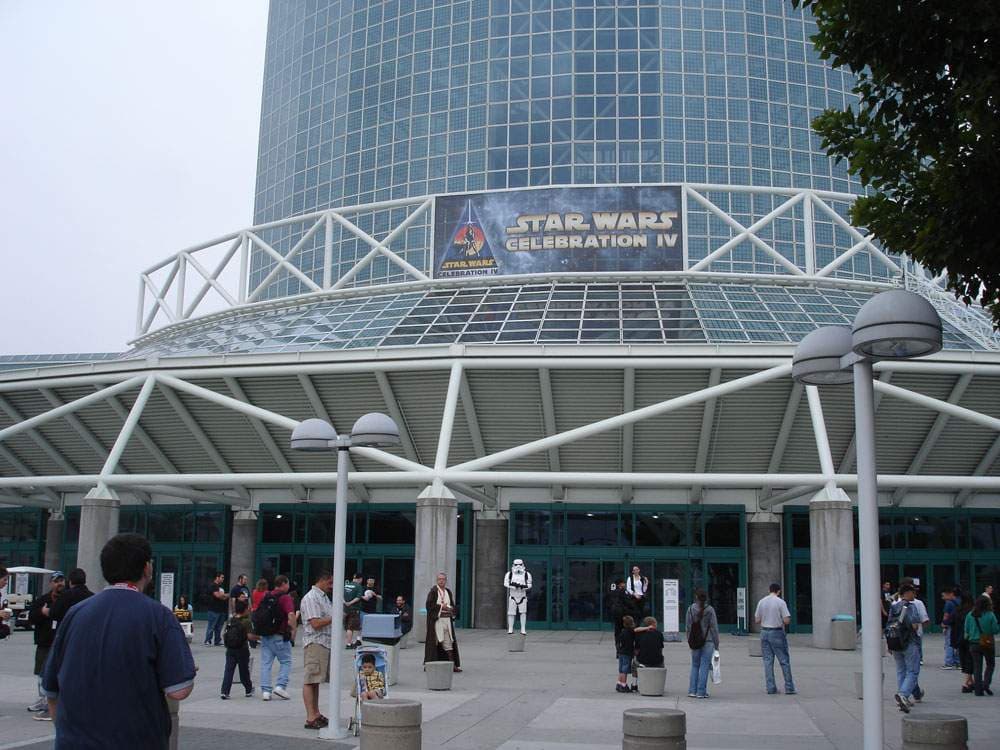 ガラス張りの円筒形の建物の下に「STAR WARS CELEBRATION IV」の大型バナーが掲げられ、白い鉄骨の庇の下の入口前を来場者が歩いている