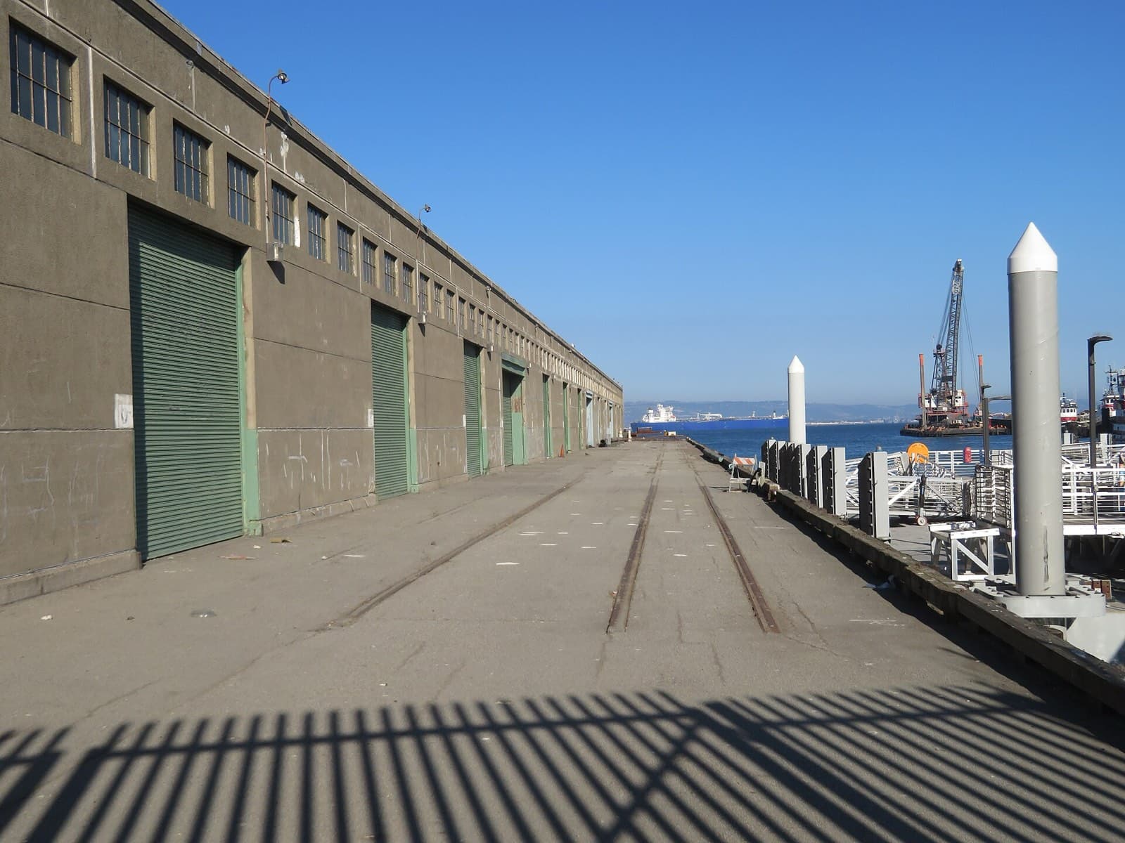 Pier 48南側の岸壁。左手に倉庫の外壁と緑色のカーゴドアが等間隔に並び、路面には貨物線のレールが2本残っている。右手は湾で、対岸に貨物船と起重機船が見える