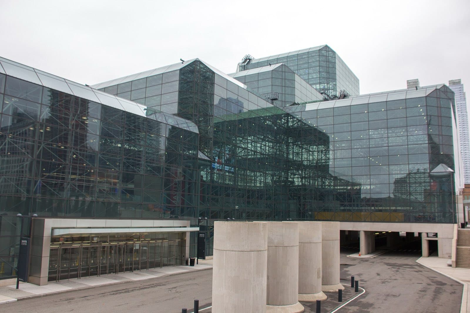 Javits Centerを車寄せ側から見た外観。ガラス張りの棟が階段状に重なり、左手には自動ドアの並ぶ来場者用の入口、右手には建物の下をくぐる車道の入口が見えます
