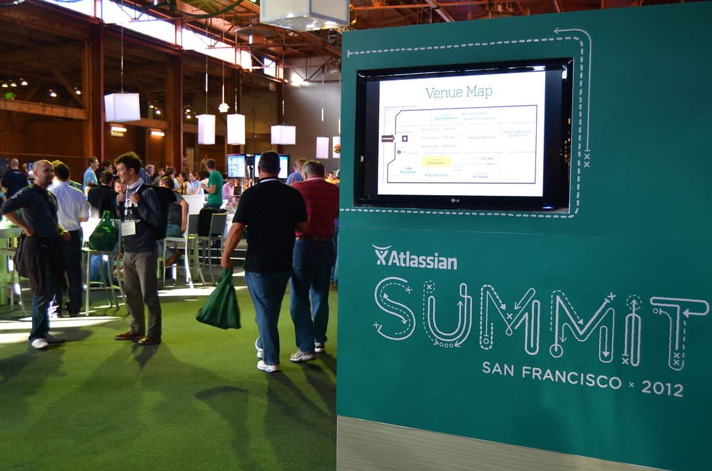 会場に立つ緑色の案内サイン。上部のモニターに会場マップが表示され、下部にAtlassian SummitとSAN FRANCISCO 2012のロゴが白抜きで記されている。左奥では参加者が歩いている