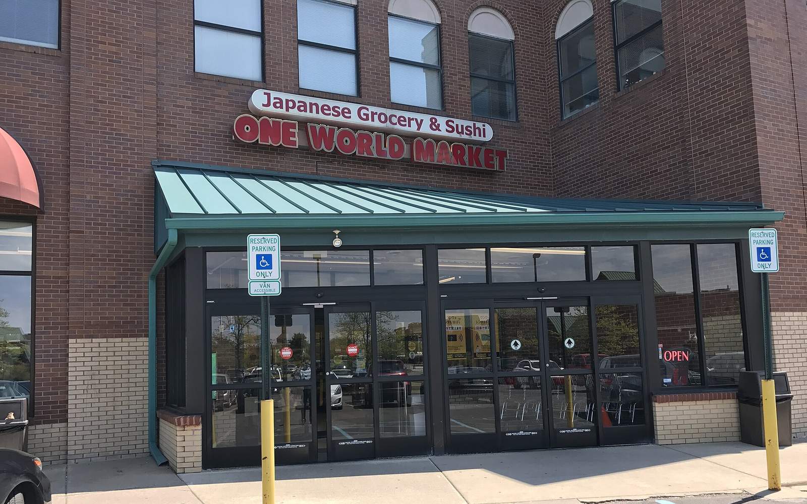 レンガ造りの建物の一階にある日本食品店の入口。「Japanese Grocery & Sushi ONE WORLD MARKET」と書かれた赤い看板と、緑色の庇、自動ドアが写っている外観写真
