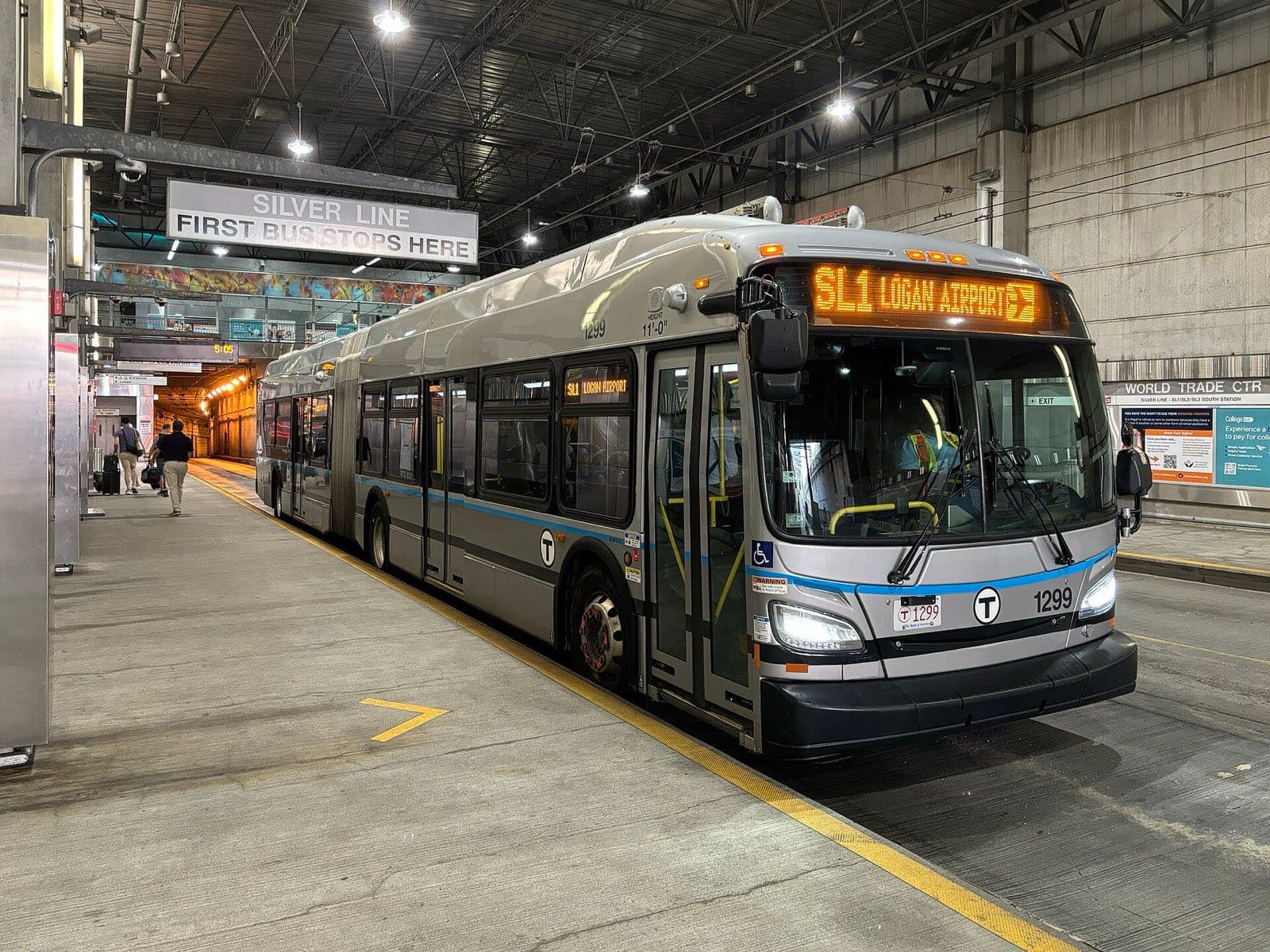 地下のバス乗り場に停車する銀色の連節バス。行き先表示にSL1 Logan Airportと出ており、奥をスーツケースを引いた乗客が歩いている
