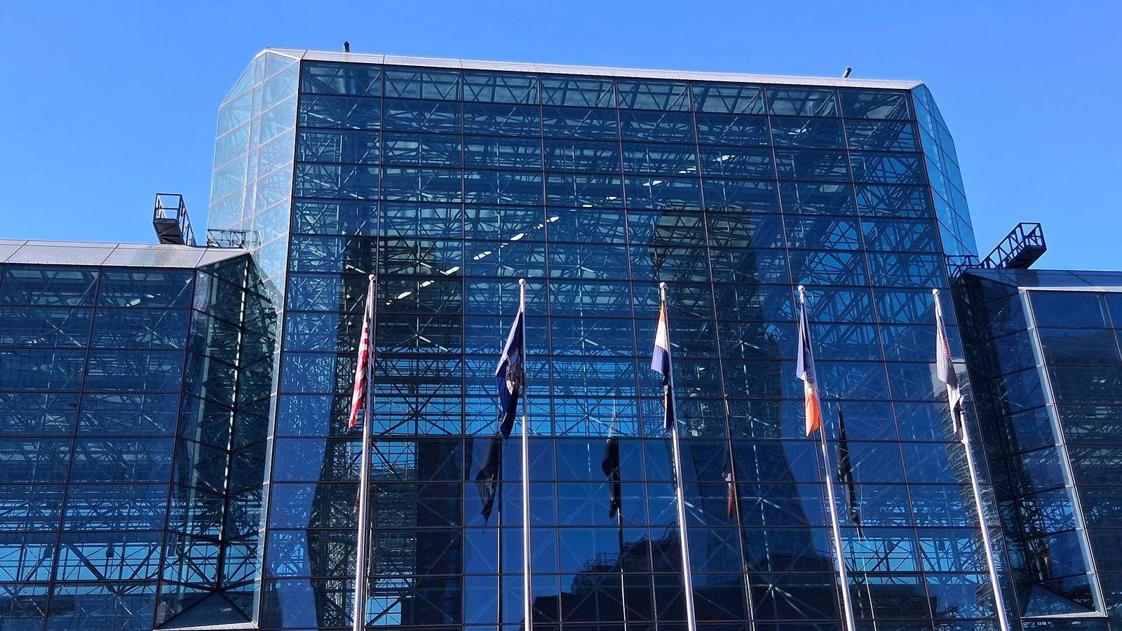 青空を背景にした、ガラス張りのJacob K. Javits Convention Centerの正面外観。中央の高い箱型の棟から左右に低い展示棟が伸び、手前に6本の旗竿が並んでいます