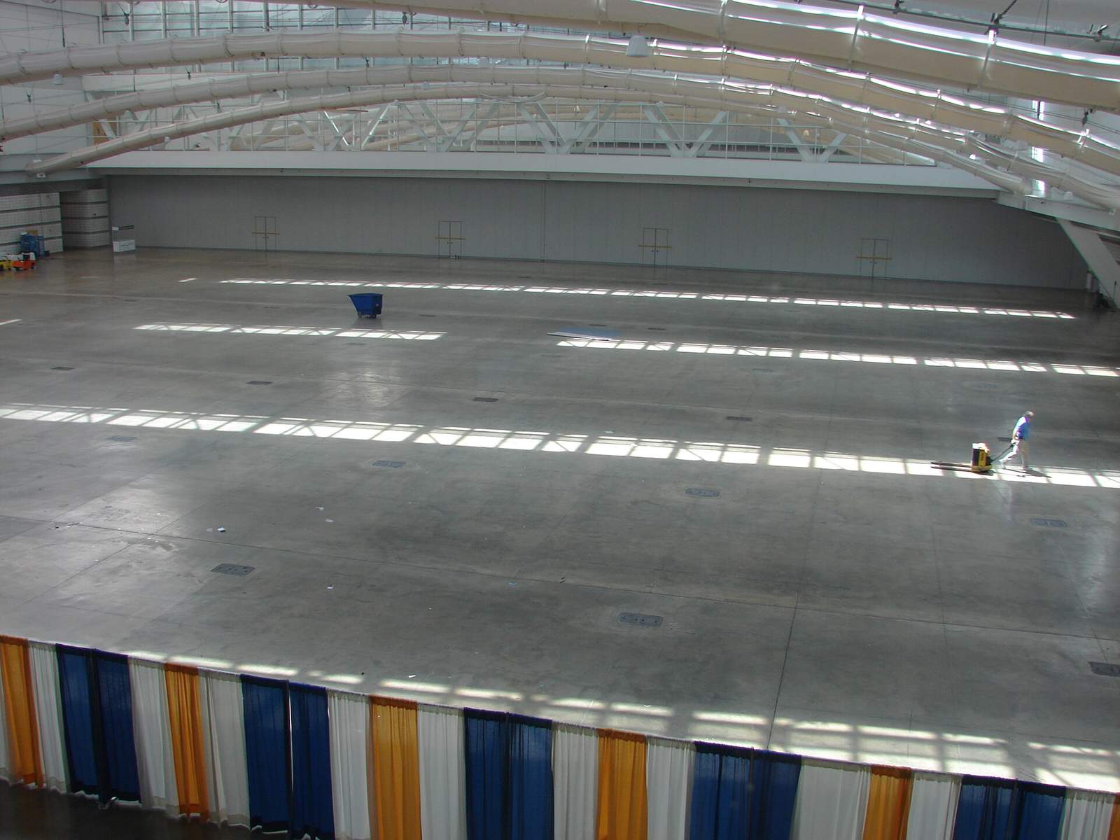 設営前で何もない展示ホールの床。コンクリートの床面に天窓からの光が縞状に落ち、遠くにハンドリフトを引く作業員が1人だけ見える