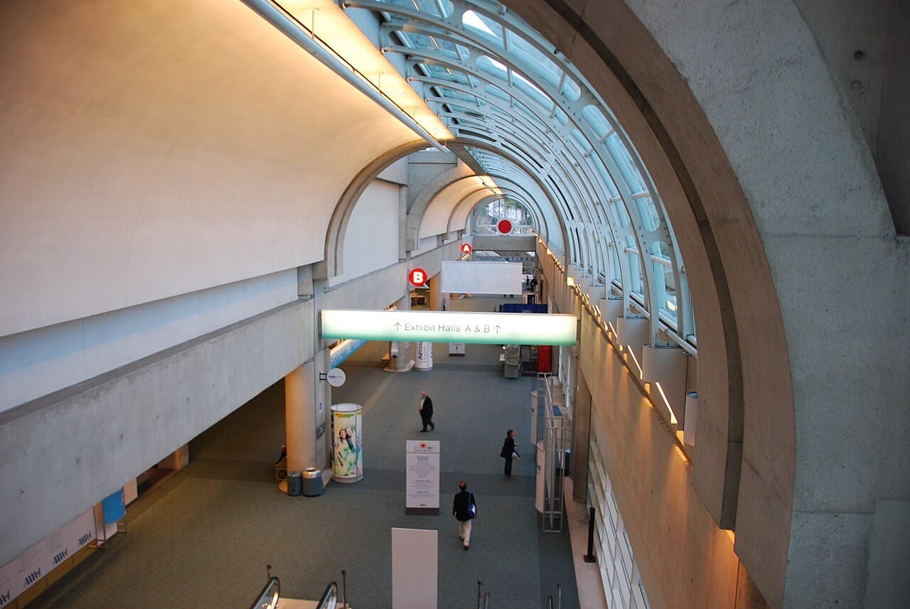San Diego Convention Centerの上層コンコース。かまぼこ型のガラス天井の下に「Exhibit Halls A & B」と光る案内サインが下がり、下階へのエスカレーターが伸びる