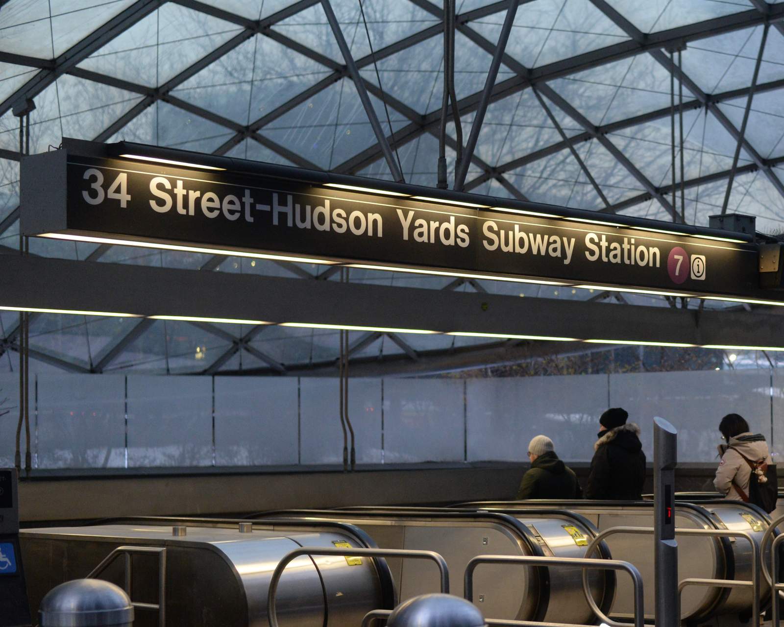 ガラスの曲面屋根に覆われた地下鉄駅の入口。「34 Street-Hudson Yards Subway Station」と7系統の丸印が入った照明看板の下に地下へ下るエスカレーターが並び、厚手のコートを着た数人が乗っています